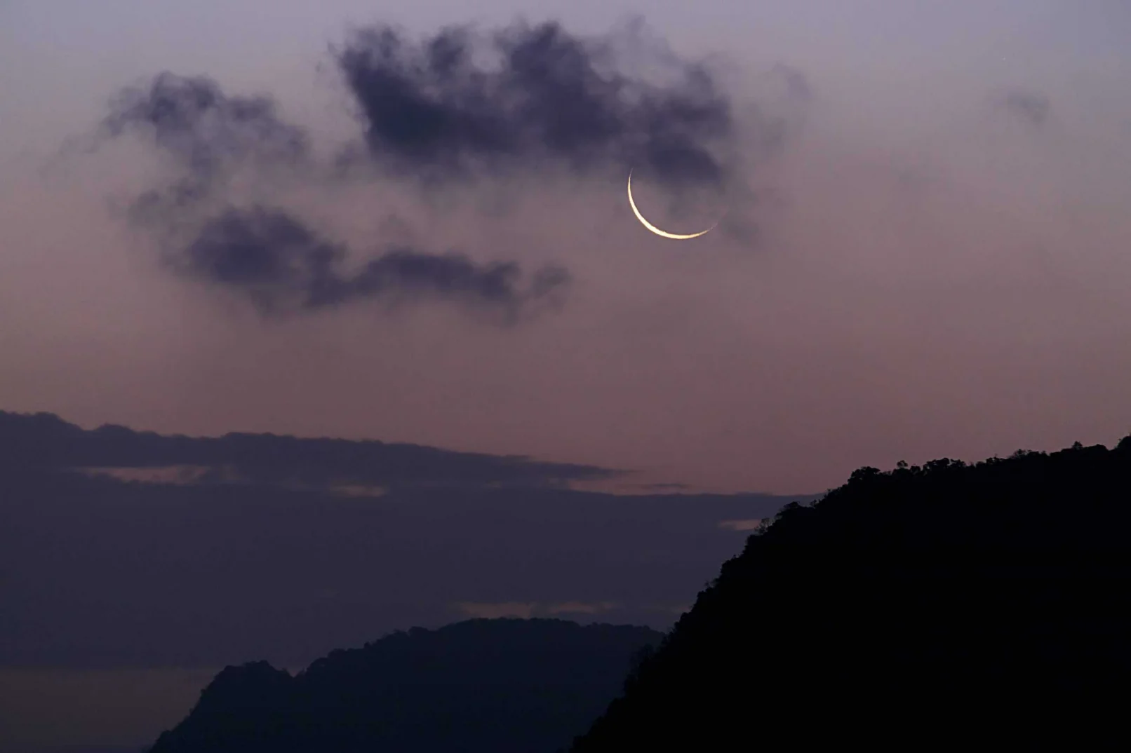 A crescent moon rises over silhouetted mountain peaks at dusk, evoking stillness and contemplative peace.